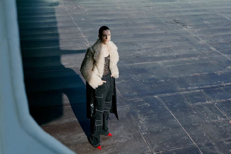 Adult fashion model poses in an outdoor setting, wearing a fur coat, black pants, and red heels, standing on a concrete surface.