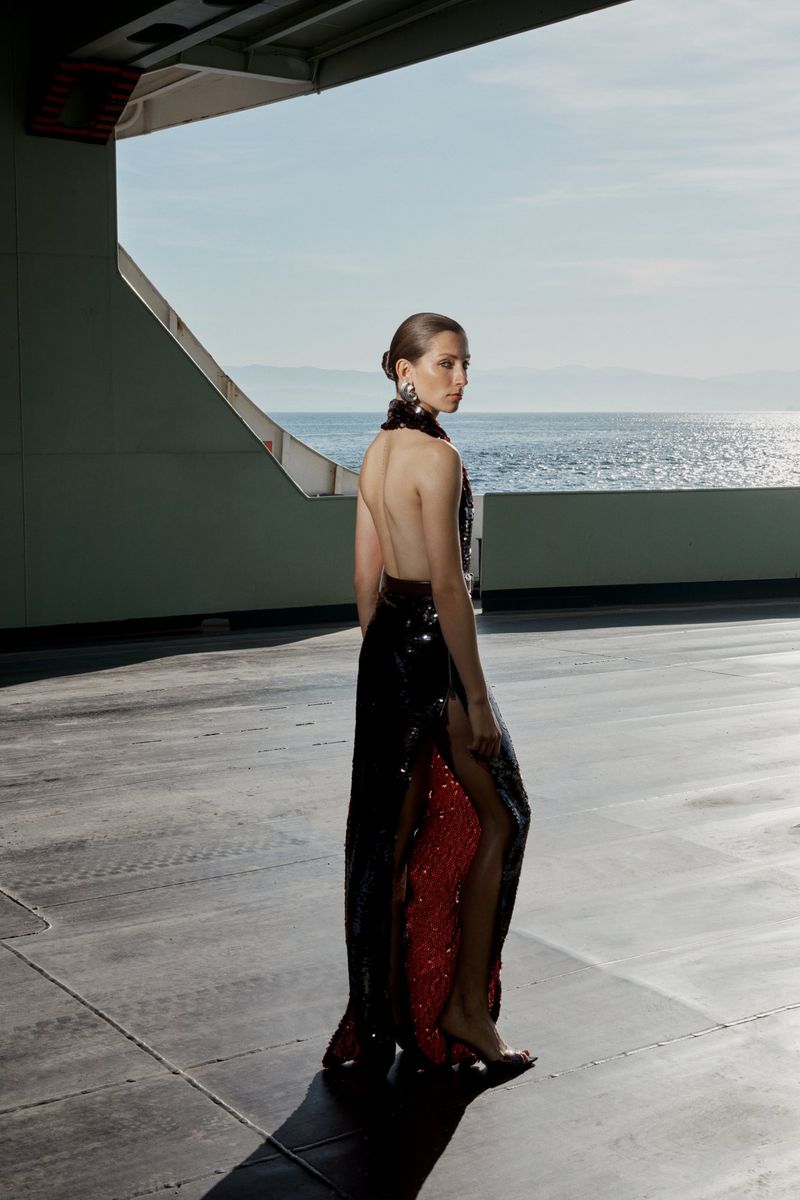 Elegant woman in a backless sequin dress stands against the ocean in a high-fashion editorial shot, showcasing her unique style.