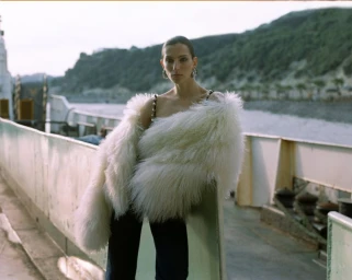 Model wearing a fluffy white fur stole and dark trousers leaning against a ferry railing with a coastal landscape behind.