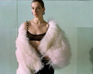 Model wearing a fluffy white faux fur coat and black lace bralette against a muted green wall for ILA fall winter collection.