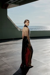 Model in a black sequined evening dress with a high slit standing on a waterfront deck under a large architectural overhang.