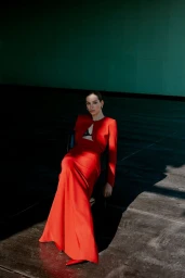 Model wearing a vibrant red dress seated on a chair against a dark interior setting with a green upper wall.