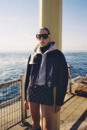 Model wearing dark sunglasses, hooded jacket, and studded miniskirt standing outdoors near a coastal railing