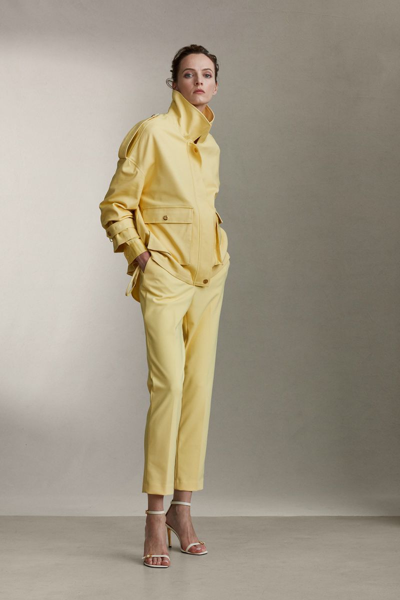 A slender woman poses in a studio wearing a yellow jacket and matching straight-leg pants with white heels and a neutral background.