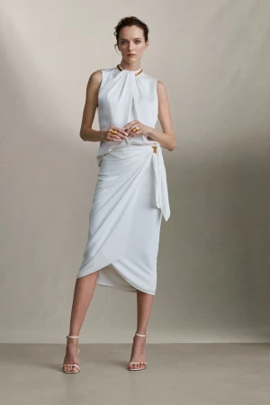 Woman in an elegant white sleeveless top and wrap skirt with gold accents stands in a studio with neutral colors, posing for fashion editorial.