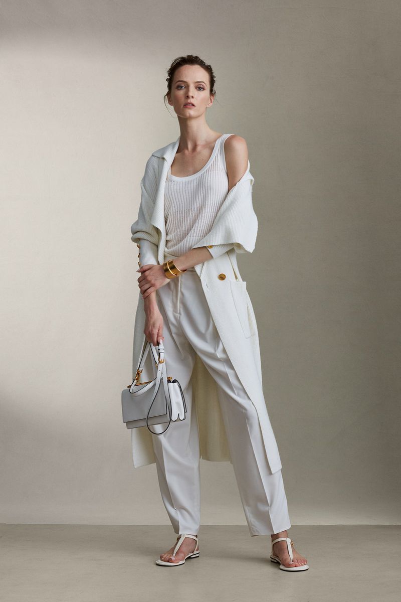 A woman in a white tank top, trousers, and trench coat poses with a small white purse against a neutral background in a fashion shot.
