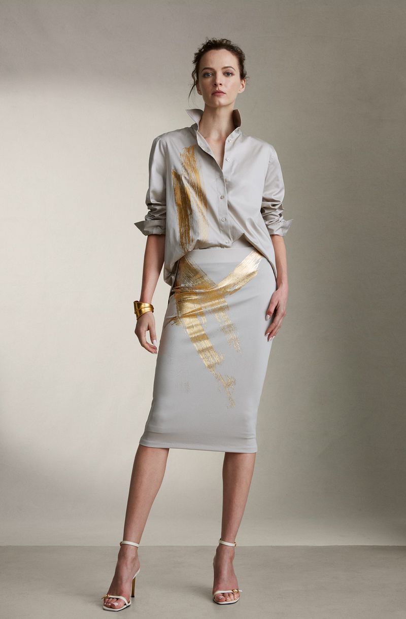 Fashion model poses in a grey satin blouse and matching pencil skirt with gold accents, paired with high heels in a studio setting.