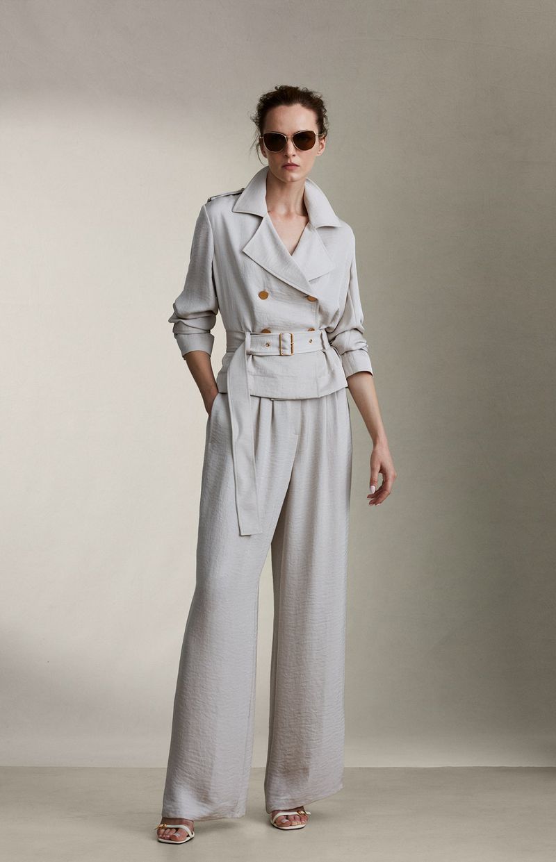 A woman in a grey suit, posing in a photo studio, wearing sunglasses and sandals. The suit is belted and has wide legs.