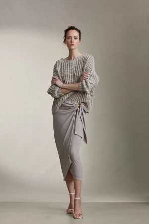 Model standing with arms crossed wearing a textured grey knit sweater and draped skirt against a seamless studio wall