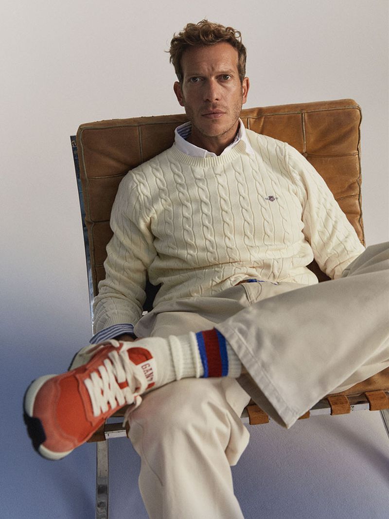 Man in off-white cable-knit sweater sitting in leather sling chair in a studio, wearing casual pants and orange sneakers.