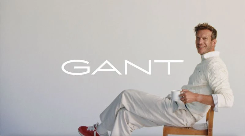 An adult male in a casual outfit sits on a wooden chair, holding a mug, with the 'GANT' logo in the background against a neutral background.