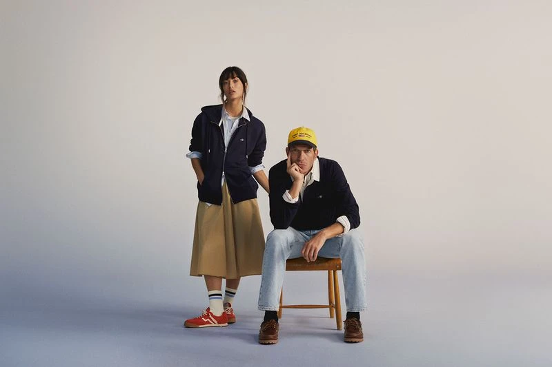 A man and woman pose in a bright studio. The man sits on a wooden chair in jeans, and a navy sweater. The woman stands in a skirt and jacket.