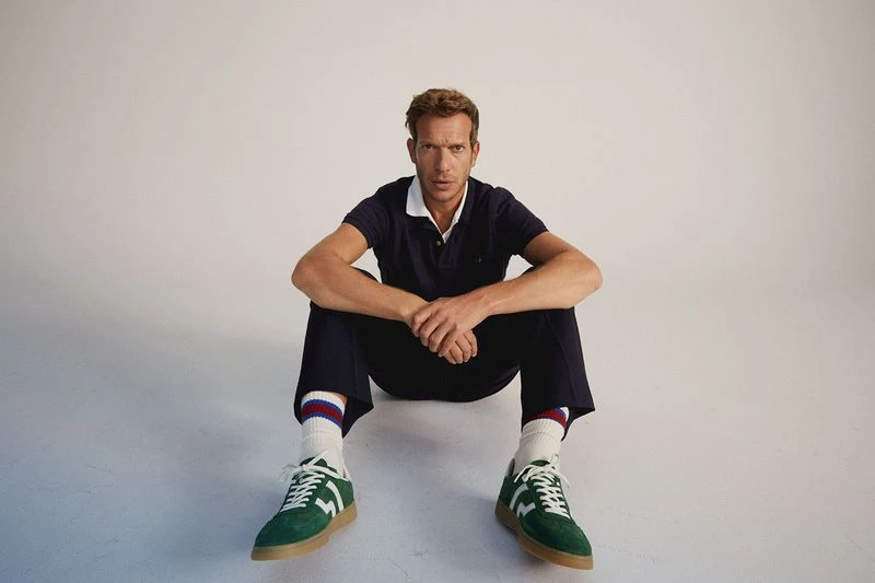 Man sits on floor wearing a blue polo shirt, dress pants, crew socks with stripes, and green sneakers against a white backdrop.