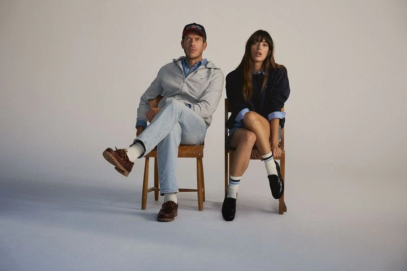 A man and woman sit on wooden chairs against a neutral backdrop, showcasing preppy fashion with a baseball cap, sweater, and loafers.