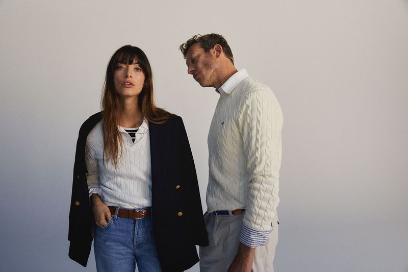 A fashionable couple poses in front of a blank backdrop, wearing matching knit sweaters and denim jeans in a studio setting.