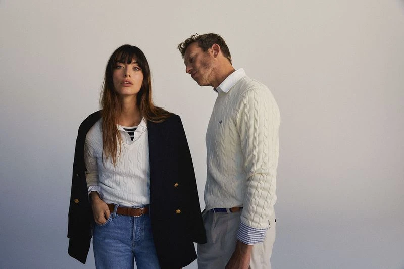 A fashionable couple poses in front of a blank backdrop, wearing matching knit sweaters and denim jeans in a studio setting.