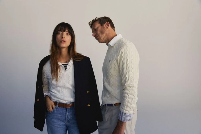 A fashionable couple poses in front of a blank backdrop, wearing matching knit sweaters and denim jeans in a studio setting.