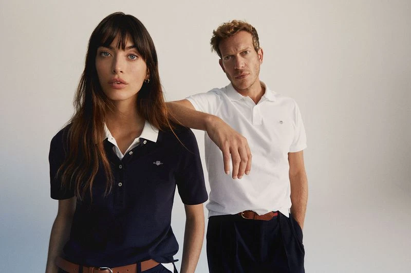 A young adult woman and man pose indoors wearing polo shirts and trousers, in a brightly lit studio. Fashion shot, neutral background.