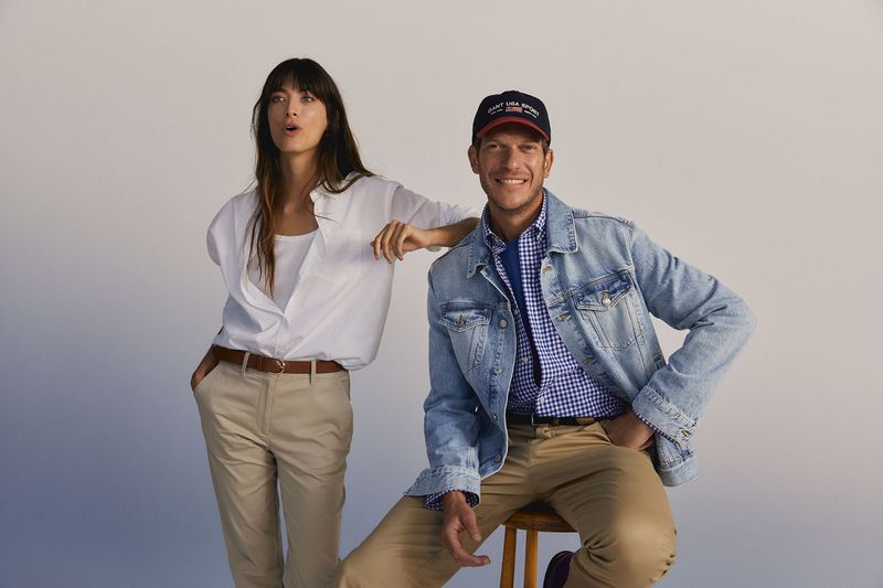 A fashionable couple, a woman in a white shirt and a man in a denim jacket, poses in a photo studio setting with a neutral background.