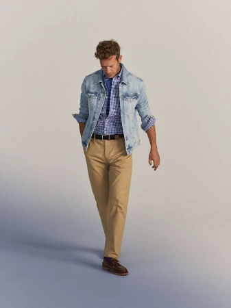 Man walks in a light denim jacket, blue gingham shirt, and khaki pants in a studio setting, presenting a casual and stylish look.