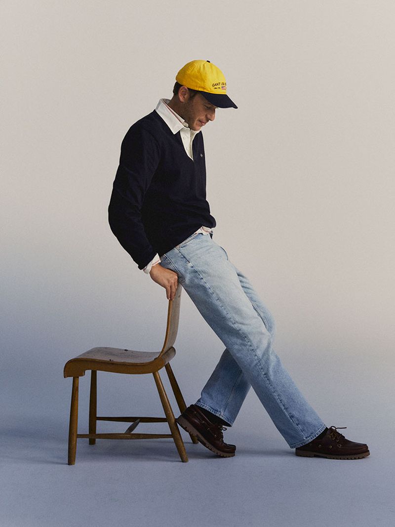 Man in blue sweater, jeans, and yellow cap leans on a wooden chair in a studio with soft, neutral lighting, fashion pose.