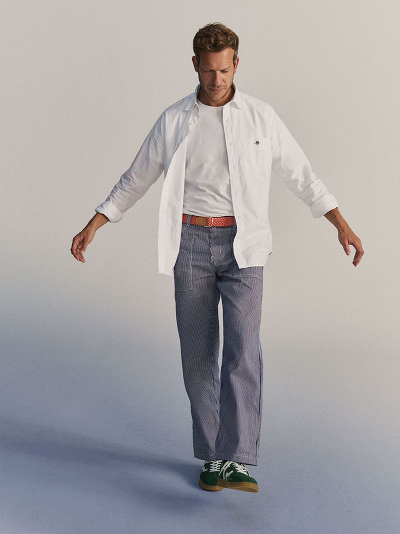 A blond male model wearing a white button-down shirt, striped pants, and green sneakers stands in a neutral studio environment.