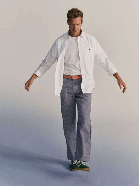 A blond male model wearing a white button-down shirt, striped pants, and green sneakers stands in a neutral studio environment.