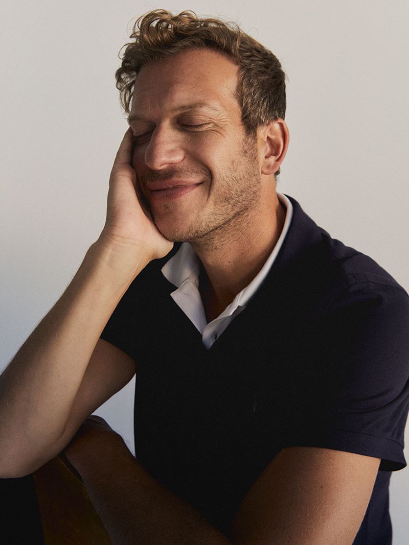 Portrait of an adult male with curly hair wearing a polo shirt, smiling with his eyes closed and resting his head on his hand in a studio.