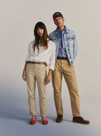A stylish couple poses in a photo studio. The woman wears a white shirt and tan pants, while the man sports a denim jacket and baseball cap.