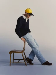 Man wearing a yellow cap, dark navy polo shirt, and light blue jeans leaning against a wooden chair.