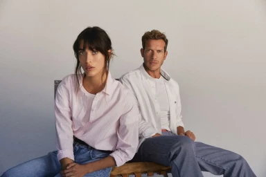 A woman in a pink striped shirt and a man in a white shirt seated together against a plain neutral background for Gant S/S 25.