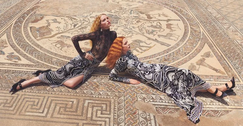 Two women in dresses with a black and white swirl pattern lay on a mosaic floor in the daylight, posing for a fashion shoot.