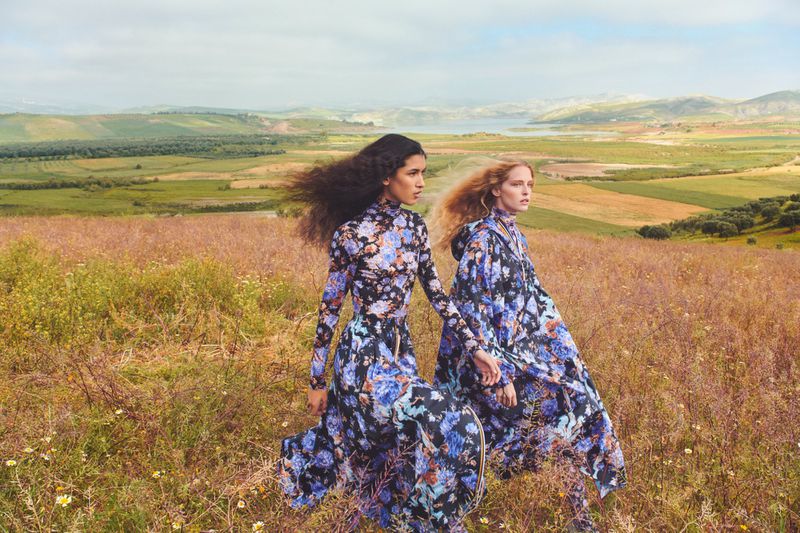 Two fashion models wearing floral dresses walk through a golden field. Landscape features green hills and a lake in the distance.