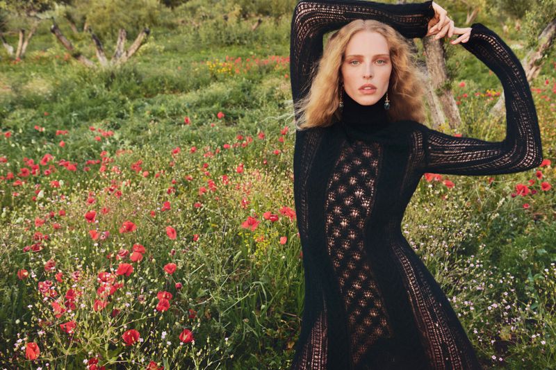 Woman in black turtleneck dress poses in sunlit field with red flowers and olive trees, capturing a natural, elegant fashion moment.