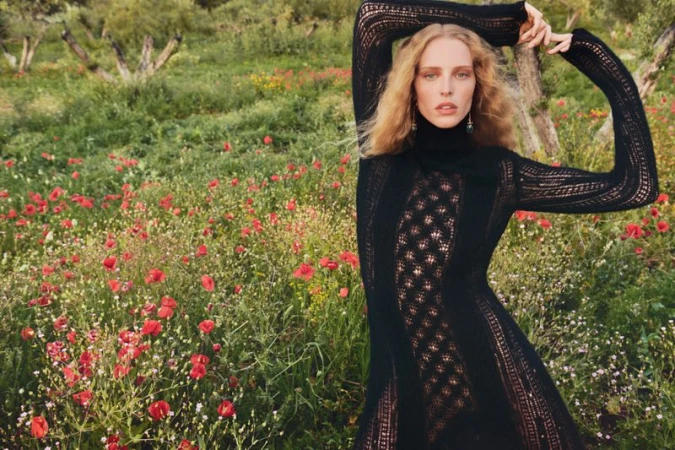 Woman in black turtleneck dress poses in sunlit field with red flowers and olive trees, capturing a natural, elegant fashion moment.