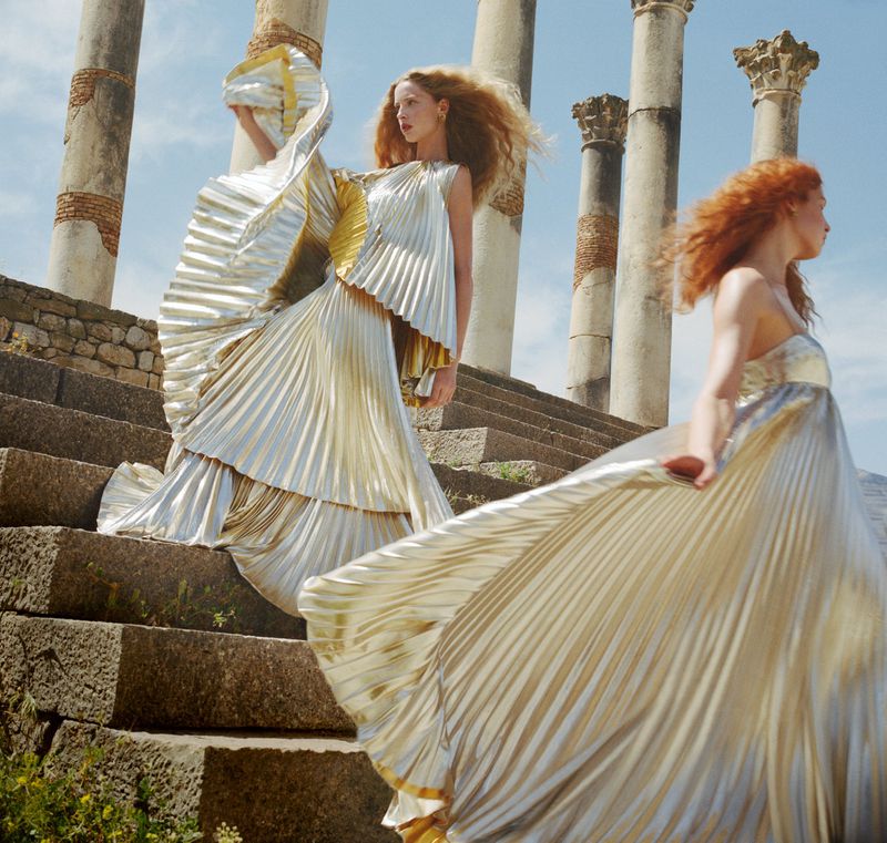 Two women in elegant gold pleated gowns pose on stone steps near ancient columns under a blue sky. Fashion editorial photography.