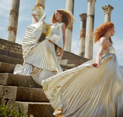Two women in elegant gold pleated gowns pose on stone steps near ancient columns under a blue sky. Fashion editorial photography.