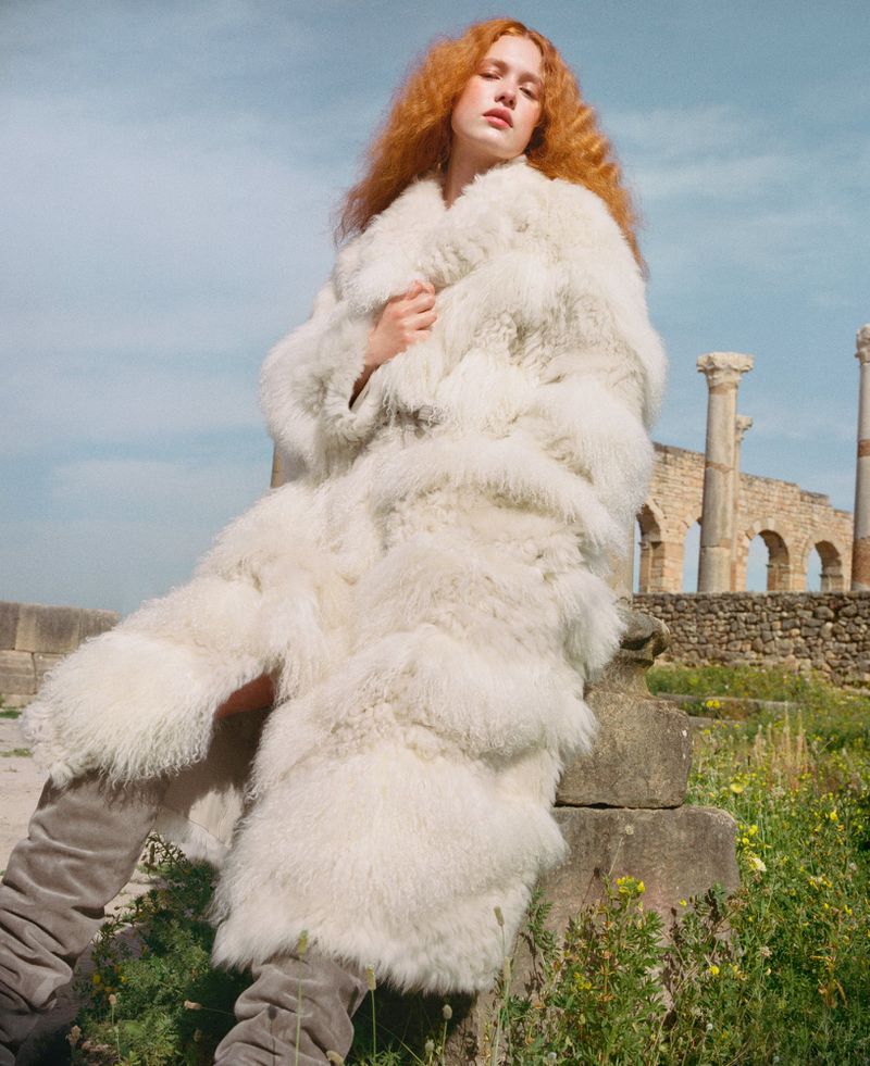Redhead woman wearing a large white fur coat posing in a field with Roman ruins on a sunny day, fashion editorial shot.