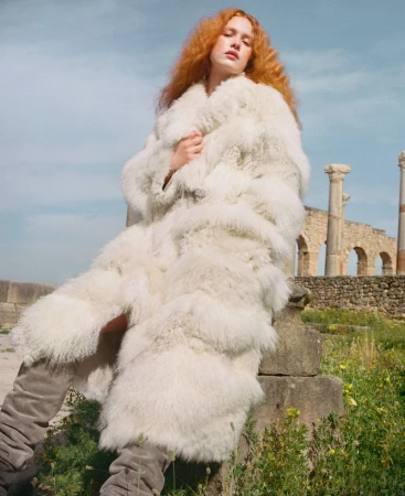 Redhead woman wearing a large white fur coat posing in a field with Roman ruins on a sunny day, fashion editorial shot.