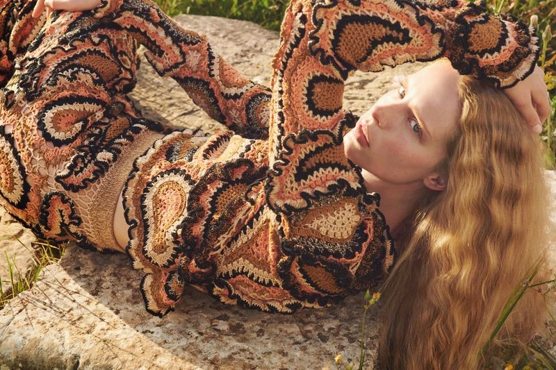 Woman in crochet dress reclines on rock surrounded by green grass. Earthy tones and textures define a natural fashion aesthetic.