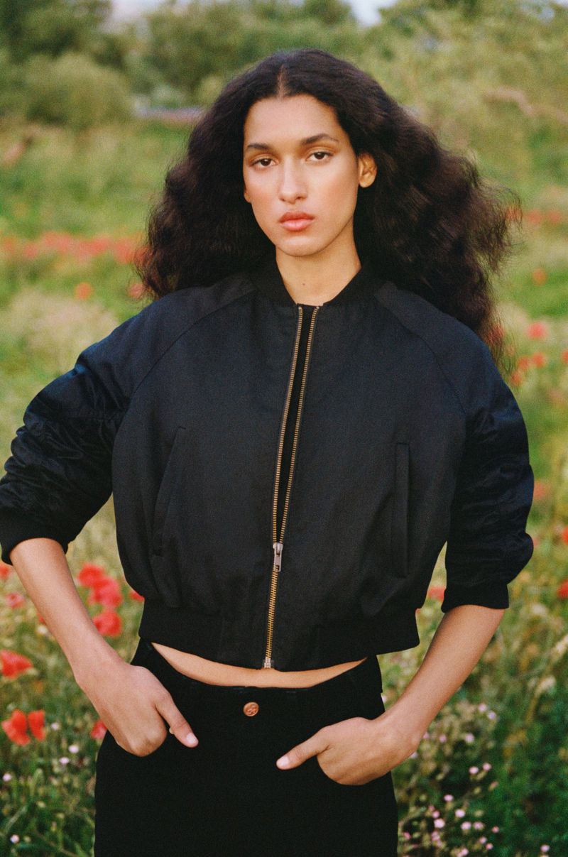 Portrait of a stylish adult female in a black bomber jacket posing outdoors in a flower-filled field with a neutral expression.