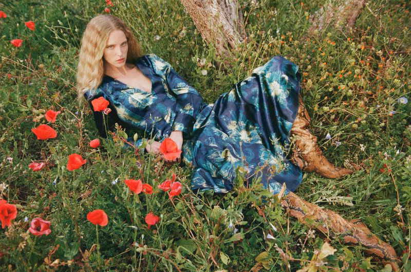 Woman in patterned dress and boots lies in a grassy field with red poppies in soft light, creating a whimsical, fashion-editorial image.