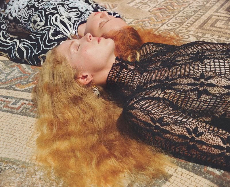 Two women lie side-by-side on a mosaic tile floor, wearing patterned clothing. One has blonde hair, the other has red hair.