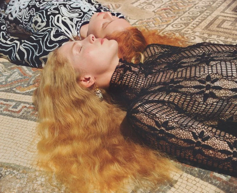 Two women lie side-by-side on a mosaic tile floor, wearing patterned clothing. One has blonde hair, the other has red hair.