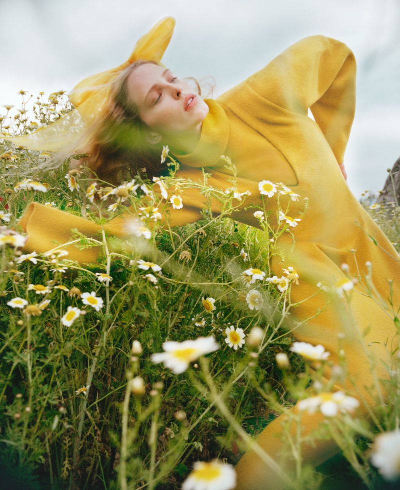 Young woman with red hair lies in a field of chamomile daisies wearing a yellow dress and headscarf, outdoor setting.