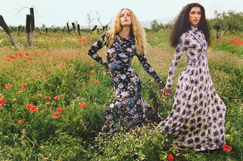 Two women in floral dresses hold hands in a wildflower field with green grass and red poppies, under an overcast sky, creating a fashion editorial.