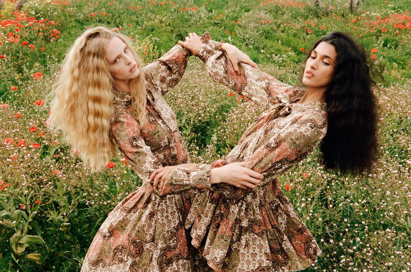 Two fair-skinned adult females in patterned dresses pose together in a flower-filled meadow setting, embracing a fashion-forward editorial style.
