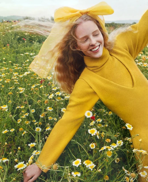 Young woman in a yellow outfit smiles radiantly in a field of white daisies, enhanced by a soft yellow bow, under diffused lighting.