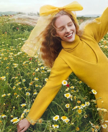 Young woman in a yellow outfit smiles radiantly in a field of white daisies, enhanced by a soft yellow bow, under diffused lighting.