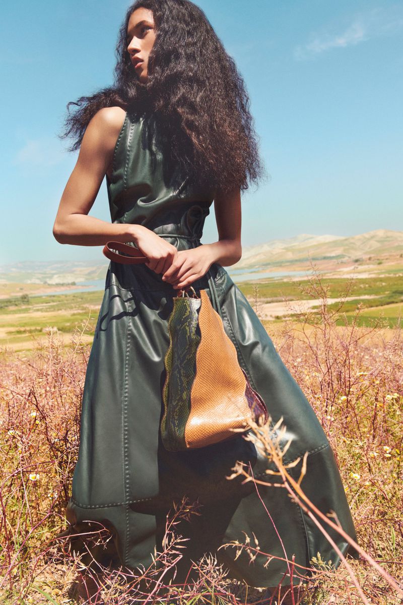 Fashion shot of woman in a green dress holding a leather bag in a sunlit, golden field against a clear blue sky. Low angle, eye-level view.